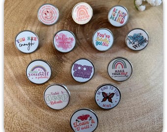 May include: A collection of round, silver-rimmed pins with various inspirational quotes and designs. The pins feature phrases like "You are enough," "Believe in yourself," and "Be kind to your mind." Some pins have rainbow and butterfly graphics.