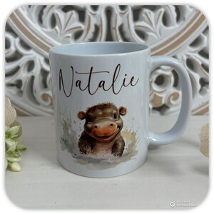 May include: White ceramic mug featuring a smiling cartoon hippo with brown and tan coloring. The name "Natalie" is written above the hippo in a brown script font. The mug has a curved handle.
