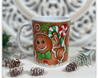 May include: A white ceramic mug with a festive gingerbread man design. The mug features a smiling gingerbread man with a green bow tie, candy canes, and a red and white swirl lollipop. The background is brown with decorative swirls.