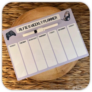 May include: A purple dry-erase weekly planner with the text "Alfie's Weekly Planner" at the top. The planner has a space for each day of the week, Monday through Sunday. A white dry-erase marker and a black and grey video game controller are included.