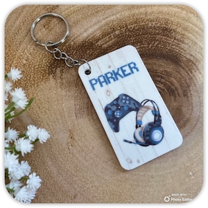 May include: A rectangular keychain with a silver keyring. The keychain features a wood-grain background with the name "PARKER" in blue block letters. Below the name are illustrations of a blue video game controller and headphones.