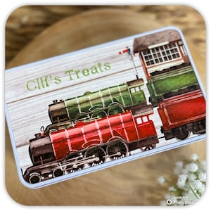 May include: A rectangular metal tin featuring a printed design of a green steam locomotive and a red steam locomotive on a light wood grain background. The text Clif's Treats is printed in green letters on the left side of the lid.