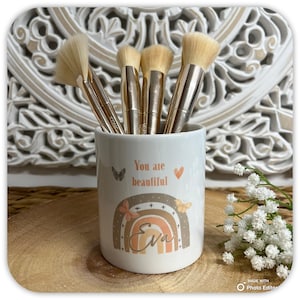 May include: A white ceramic makeup brush holder with the text "You are beautiful" and a rainbow design. The holder contains several makeup brushes with gold handles and soft bristles. The name "Eva" is written on the rainbow.