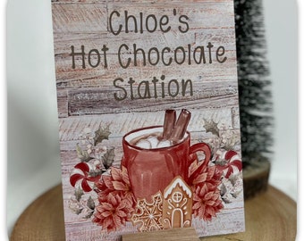 May include: A rustic wooden sign with the text "Chloe's Hot Chocolate Station" in brown lettering. The sign features a watercolor illustration of a red mug filled with hot chocolate, marshmallows, and cinnamon sticks, surrounded by poinsettias, candy canes, and a gingerbread house.