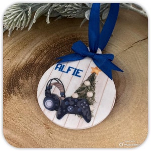 May include: A round, white, wooden Christmas ornament with a blue ribbon. The ornament features a cartoon illustration of a video game controller, headphones, and a Christmas tree with the name "Alfie" printed on it.