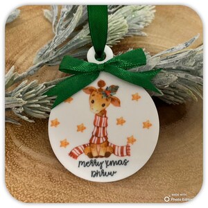May include: A round white ornament featuring a cartoon giraffe wearing a red and white striped scarf, decorated with small orange stars. The text "merry xmas" is printed in black below the giraffe, topped with a green satin ribbon bow.