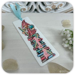May include: White bookmark with a gnome illustration wearing a pink dress and hat, holding a globe. The bookmark features pencils and books, with the text "Miss Dawn's Bookworm". A light blue ribbon is attached.