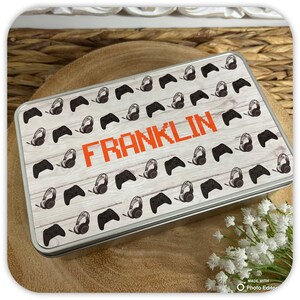 May include: A silver metal tin with a woodgrain background and a repeating pattern of black video game controllers and headphones. The name "FRANKLIN" is printed in orange pixelated letters on the tin.