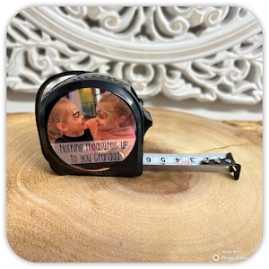 May include: Black tape measure with a photo of two children and the text "Nothing measures up to you Grandad." The tape extends to show measurements in inches. The tape measure is on a wooden surface.