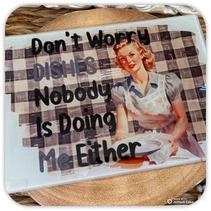 May include: A white chopping board with a black and white checked background. The text "Don't Worry Dishes Nobody Is Doing Me Either" is written in black on the board. A woman in a floral apron is holding a plate in front of her.