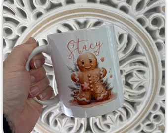 May include: White ceramic mug featuring a watercolor gingerbread man design with a star and pine accents. The name "Stacy" is written in a cursive font above the gingerbread man. The mug has a white handle.