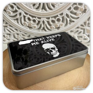 May include: A rectangular metal tin with a black lid featuring a white skull and the text "THAT KEEPS ME ALIVE". The tin sits on a wooden surface, with a decorative white background.