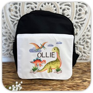 May include: Black backpack with a white front pocket. The pocket displays a colourful dinosaur illustration, including a green Brontosaurus, an orange Stegosaurus, and a Pterodactyl, with the name "OLLIE".