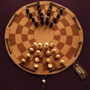 Puede incluir: Un tablero de ajedrez bizantino circular de material marrón y beige, con piezas de ajedrez negras y blancas dispuestas en el tablero. El tablero tiene una etiqueta que dice "Byzantine Chess".