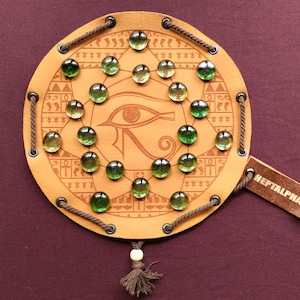 Puede incluir: Un juego de mesa redondo de cuero marrón claro con canicas de vidrio verde. El tablero presenta un símbolo de ojo y grabados decorativos. Una etiqueta de cuero dice "REPTALPHA". El juego está ensartado con cuerda y tiene una borla.
