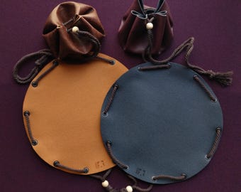 May include: Leather pouches and round mats in shades of brown and blue. The pouches have drawstring closures with wooden beads. The mats have a drawstring around the edge. The mats have the initials "IF3" stamped on them. The background is a dark purple.