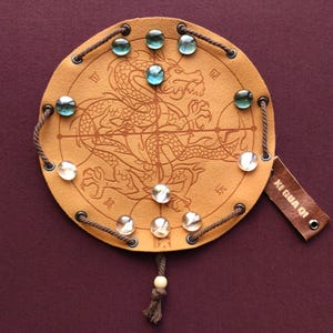 May include: A tan leather game board with a dragon design and a string border. The board has nine holes with glass marbles in each hole. The board has Chinese characters on it and a tag that says "KI GWA OI".