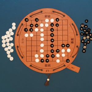 Può includere: Una tavola da gioco Go rotonda in pelle color cuoio con motivo a griglia, dettagli in corda marrone scuro e caratteri asiatici. La tavola presenta pedine bianche e nere, con pietre bianche sparse a sinistra e pietre nere a destra.