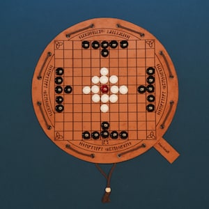 Op de afbeelding: Een rond bordspel van beige leer met een rasterpatroon, gegraveerde runenteksten en decoratieve knoopmotieven. Het bord is gevuld met zwarte, witte en één rode bolvormige speelstukken. Een leren koord loopt langs de rand van het bord.