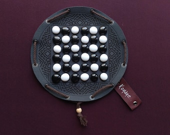 Op de afbeelding: Een rond, grijs leren bordspel met een geometrisch ontwerp en een raster van zwarte en witte spelstukken. Het bord heeft een bruin trekkoord en een label met de tekst "Koyane".
