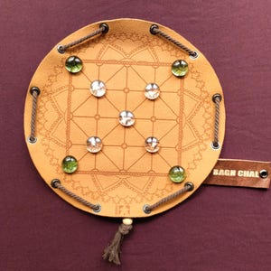 Puede incluir: Un tablero de juego de Bagh Chal de cuero con un fondo marrón y un diseño de mandala decorativo. El tablero tiene nueve cuadrados y está rodeado por un borde de cuerda. Hay ocho canicas de vidrio en el tablero, seis blancas y dos verdes.