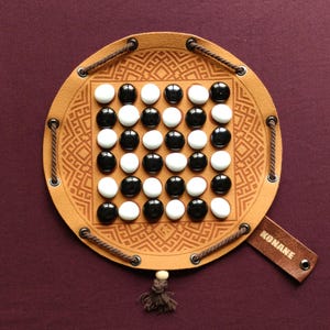Puede incluir: Un tablero de juego de cuero marrón con un patrón blanco y negro y piezas de juego blancas y negras. El tablero tiene una correa de cuero con la palabra "KONANE" impresa en ella.