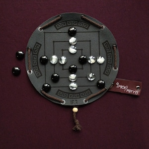 Puede incluir: Un juego de mesa Nine Men's Morris hecho de material gris oscuro con un diseño geométrico. El tablero tiene piezas de juego negras y transparentes. Una etiqueta de cuero marrón con el texto "9 men's morris" está adjunta.