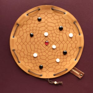 Puede incluir: Un juego de mesa de cuero marrón con un diseño circular y una red de círculos interconectados. El tablero presenta piezas de juego negras, blancas y rojas. Se adjunta una etiqueta de cuero con el nombre "FIDCHELL". El juego está sobre un fondo rojo oscuro.
