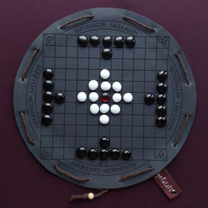 Puede incluir: Un juego de mesa redondo de cuero gris oscuro con un patrón de cuadrícula. Las piezas del juego son negras, blancas y una roja. El tablero tiene lazos de cuerda marrón y una etiqueta de cuero. El juego está sobre un fondo morado oscuro.