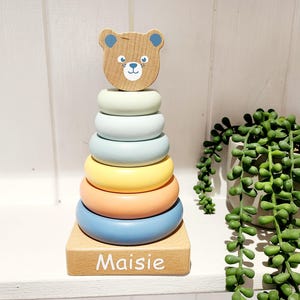 May include: A wooden stacking toy with a bear face on top. The rings are in shades of blue, yellow, and orange. The base of the toy is a wooden block with the name "Maisie" written on it.