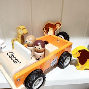 May include: A wooden toy jeep with a name tag that says "Oscar" on the front. The jeep is orange and has four black wheels. There are two wooden figures in the jeep, a giraffe, a monkey, a zebra, and a lion.