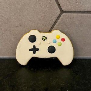 May include: A biscuit shaped like a video game controller with off-white icing. It features a black directional pad, a black circular button, and smaller yellow, blue, red, and green circular accents arranged on the surface of the controller.