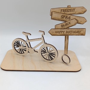 Könnte beinhalten: Eine Holzfigur eines Fahrrads mit einem Wegweiser. Der Wegweiser hat Pfeile mit den Wörtern "FREIZEIT", "SPAB", "GLÜCK" und "HAPPY BIRTHDAY". Das Fahrrad und der Wegweiser stehen auf einer Holzbasis.