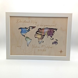 May include: A rectangular white frame containing a light wood panel with a world map cutout. The map reveals colorful paper currency behind it. Text reads: Der sch&ouml;nste Weg ist der gemeinsame!, Nadine & Stefan, and 25.05.2025, with a compass.