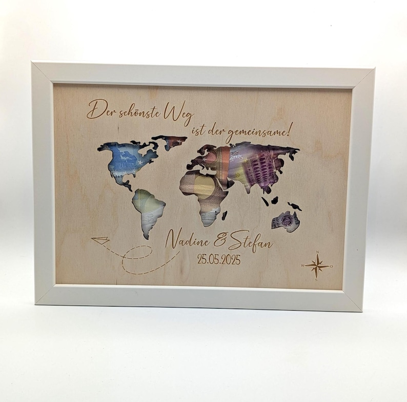 May include: A rectangular white frame containing a light wood panel with a world map cutout. The map reveals colorful paper currency behind it. Text reads: Der sch&ouml;nste Weg ist der gemeinsame!, Nadine & Stefan, and 25.05.2025, with a compass.