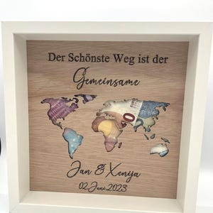 Op de afbeelding: Een houten wereldkaart in een frame met uitgesneden continenten, gevuld met kleurrijk geld. De tekst "Der Schönste Weg ist der Gemeinsame" staat boven de kaart, met "Jan & Xeniya" en "02 Juni 2023" eronder.