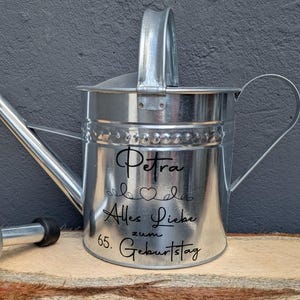 Könnte beinhalten: Eine Gießkanne aus verzinktem Metall mit Henkel und Ausguss. Der Schriftzug "Petra", die Worte "Alles Liebe zum 65. Geburtstag" und ein Herzdesign sind in schwarzer Schrift angebracht. Für die Gartenarbeit.