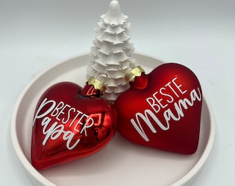 May include: Two red heart-shaped Christmas ornaments with white lettering. One ornament says "Best Papa" and the other says "Beste Mama".