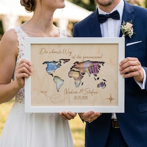 May include: A rectangular white wooden frame containing a light-colored wood panel with a laser-cut world map. The text reads Der sch&ouml;nste Weg ist der gemeinsame!, Nadine & Stefan, and 25.05.2025. A small paper airplane icon with a dashed flight path is visible.