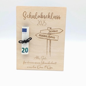 Op de afbeelding: Houten plaat met de gegraveerde tekst "Schulabschluss 2025". Een opgerolde twintig euro biljet is vastgebonden met een zwart lint. De plaat heeft ook een wegwijzer met pijlen die wijzen naar "SCHULE" en "AUSBILDUNG".
