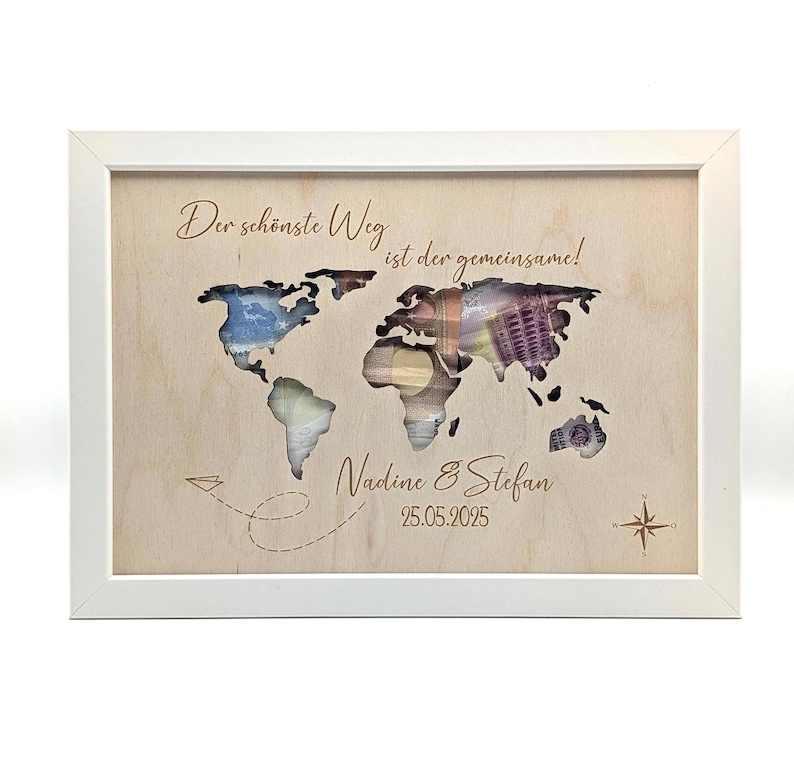 May include: A rectangular white frame containing a light wood panel with a world map cutout. Behind the cutout are folded banknotes. The text reads: Der sch&ouml;nste Weg ist der gemeinsame!, Nadine & Stefan, and 25.05.2025. A paper plane and compass are engraved.