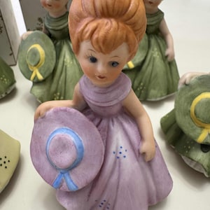 May include: A collection of ceramic figurines of young girls. The central figure wears a lavender dress and holds a matching hat with a blue ribbon. Other figures wear green dresses and hold hats. The figurines have detailed facial features.