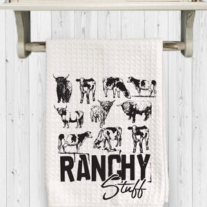 May include: White kitchen towel with a black and white illustration of cows and the text "Ranchy Stuff".