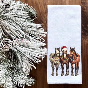 May include: White kitchen towel with a Christmas-themed design featuring three horses wearing Santa hats and Christmas lights. The horses are brown, white, and black.