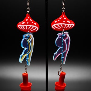 May include: A pair of red and white mushroom-shaped earrings with a blue and yellow figure hanging from each. The figures are attached to a silver chain and a red silicone earplug.