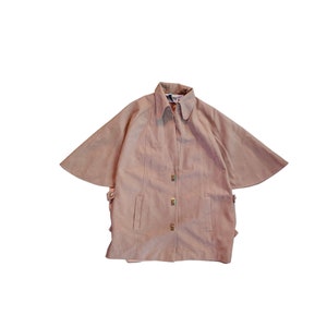 May include: A light brown cape-style jacket with a collar and three gold-coloured buttons. The jacket has a unique design with wide, flowing sleeves and two front pockets. The garment is made of a soft, suede-like material.