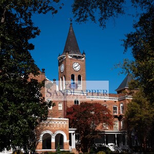May include: A brick building with a clock tower and a bell. The building has a red brick exterior and a white trim. The clock tower has a pointed roof and a bell in the top. The building is surrounded by trees and a grassy lawn.