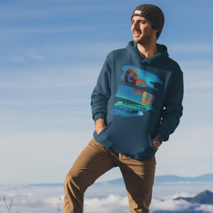 May include: A teal hoodie featuring the text "GOD IS ALWAYS HERE" in orange and blue. The hoodie has a front pocket and a drawstring hood. The person in the image is wearing a grey beanie and brown trousers.