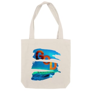May include: A cream-colored tote bag with the words "God is Always Here" in orange and blue. The text is layered over a blue and green abstract design. The bag has two cream-colored handles.