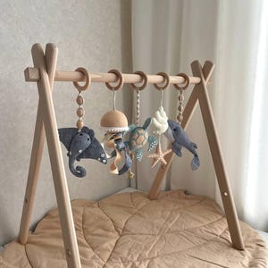 Op de afbeelding: Een houten babygym met hangende zeedierspeeltjes. De speeltjes zijn onder andere een grijze rog, een bruine paddenstoel, een blauwe schildpad, een witte zeeschelp, een zeester en een grijze walvis. De gym staat op een beige, bladvormige speelmat.